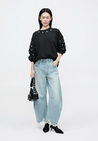 Sort sweatshirt med øjenhulsdetaljer på ærmerne, kombineret med lyseblå slidte jeans med vidde ben og en lille sort håndtaske.