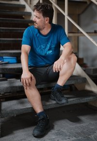 Camiseta azul de manga corta con un diseño geométrico de un oso, combinada con pantalones cortos oscuros y zapatos de senderismo negros sobre escaleras grises.