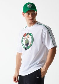 Jersey de malla de los Boston Celtics en blanco con detalles en verde. Presenta un logo serigrafiado del mascota sosteniendo un balón de baloncesto. Se combina con una gorra verde.