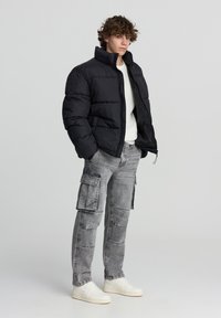 Giovane uomo che indossa una giacca a vento nera aperta, un maglione bianco, jeans cargo grigi e sneakers bianche, in piedi davanti a uno sfondo chiaro e semplice.