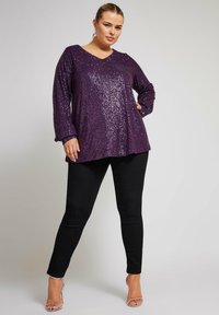 Lila paljettrik långärmad topp med V-ringning, i kombination med svarta tajta byxor. Toppens silhuett är lös och har en texturerad glans.