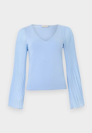 Hele sinine pikkade varrukatega särk, millel on V-kaelus ja volditud, lehvikukujulised varrukad, mis on eksponeeritud lihtsa taustaga.