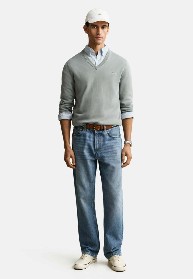 Hombre con gorra blanca, camisa de botones azul claro bajo un suéter gris de cuello en V, jeans azules, cinturón marrón, zapatillas blancas y reloj de pulsera plateado.