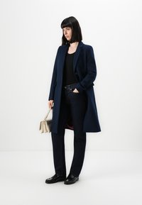 Frau mit glattem schwarzen Haar, die einen blauen Mantel, ein schwarzes Oberteil, dunkle Jeans und schwarze Schuhe trägt und eine beigefarbene Handtasche vor einem weißen Hintergrund hält.