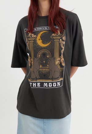 Persoon draagt een zwart oversized T-shirt met een tarotkaartontwerp waarop twee katten, zuilen, een halvemaan en de tekst "DE MAAN" te zien zijn.