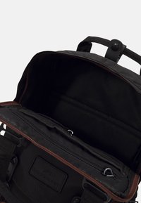 Schwarzer Stoff-Rucksack mit braunem Reißverschlussbesatz. Das Innere verfügt über eine Reißverschlusstasche, zwei offene Fächer und ein gesticktes Logoetikett.