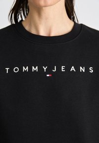 Felpa nera con collo rotondo, con il logo "TOMMYJEANS" ricamato in bianco e un piccolo accento rosso e blu. Tessuto morbido.
