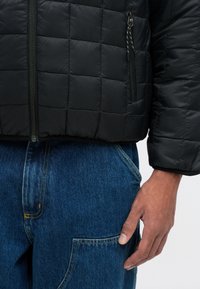 Giacca trapuntata nera con zip frontale, caratterizzata da un motivo testurizzato; indossata con pantaloni in denim blu che evidenziano una grande tasca laterale.