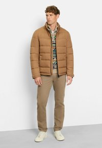 Braune Steppjacke mit gestepptem Design, Reißverschluss und hohem Kragen, getragen über einem gemusterten Pullover und kombiniert mit beigen Hosen und hellen Schuhen.