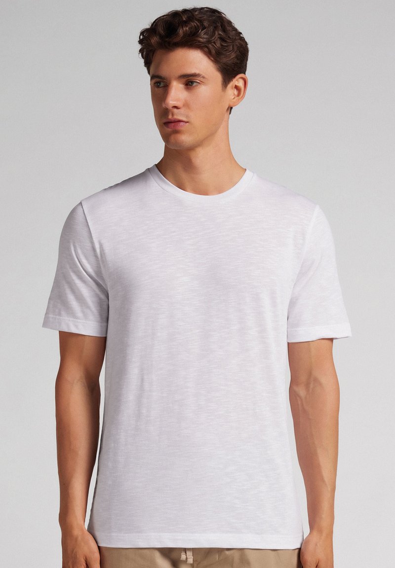 Junger Mann mit kurzen lockigen braunen Haaren, der ein schlichtes weißes kurzärmeliges T-Shirt trägt und vor einem neutralen Hintergrund steht.