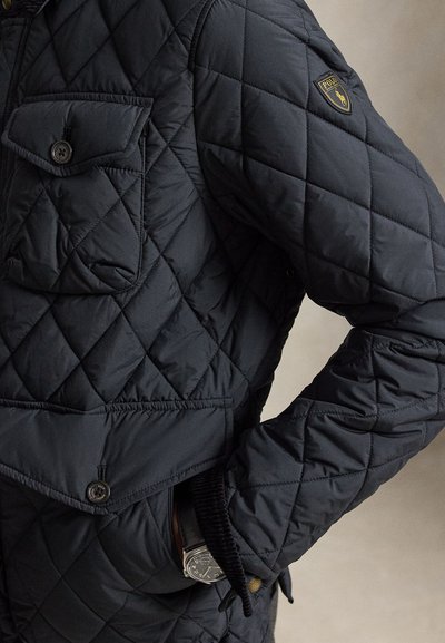 Tume tikanditega jope, millel on rinnatasku nupuga ja Polo Ralph Laureni logomärgiga varrukal, kandja käsi on küljetaskus.