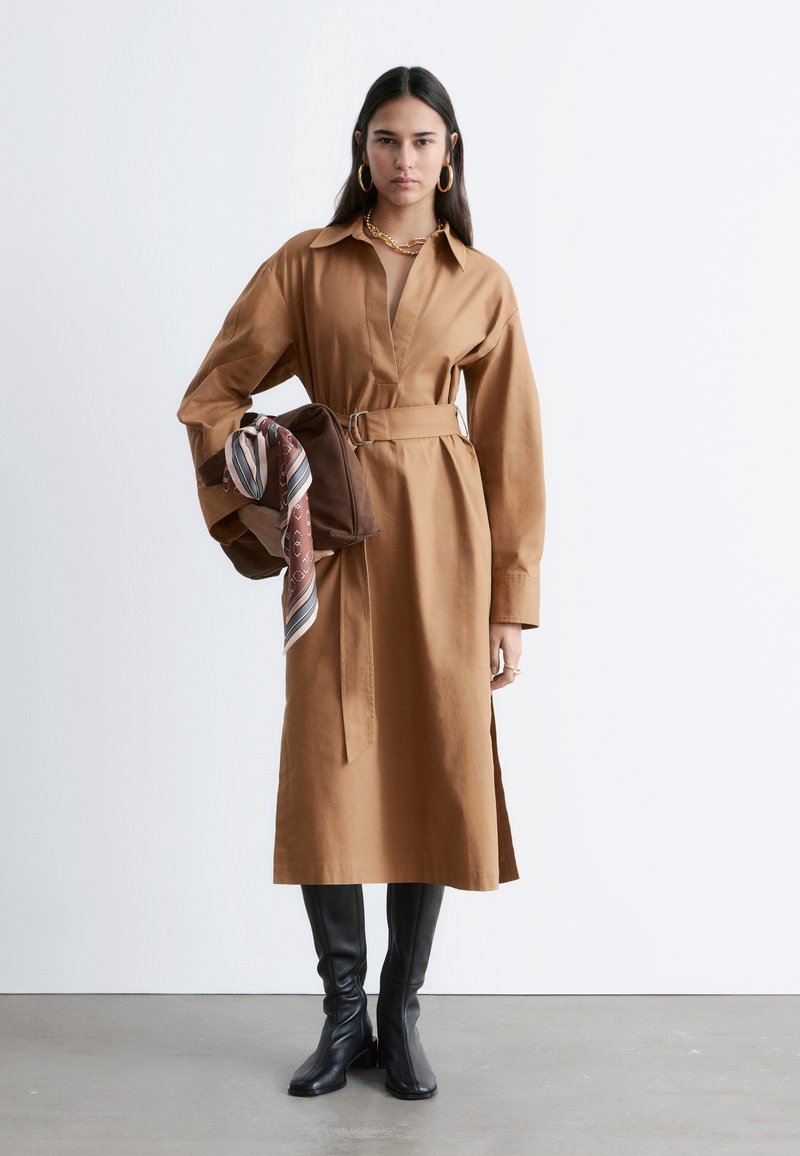 Femme portant une robe-chemise camel ceinturée et des bottes noires montantes, tenant un sac marron avec un foulard rayé attaché.