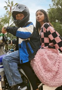 Svart og blå motorsykkeljakke, volang rosa skjørt, rutete rosa og svart genser, blå jeans, svarte sko. Motorsyklister på en motorsykkel utendørs.