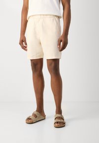 Beige linen shorts with an elastic waistband, featuring a relaxed fit and side pockets. Paired with light brown suede sandals.