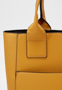 Żółta torebka typu tote z teksturowanej skóry z czarnymi detalami, z podwójnymi uchwytami i prostokątnym kształtem. Wyrazisty detal przeszycia na zewnątrz.
