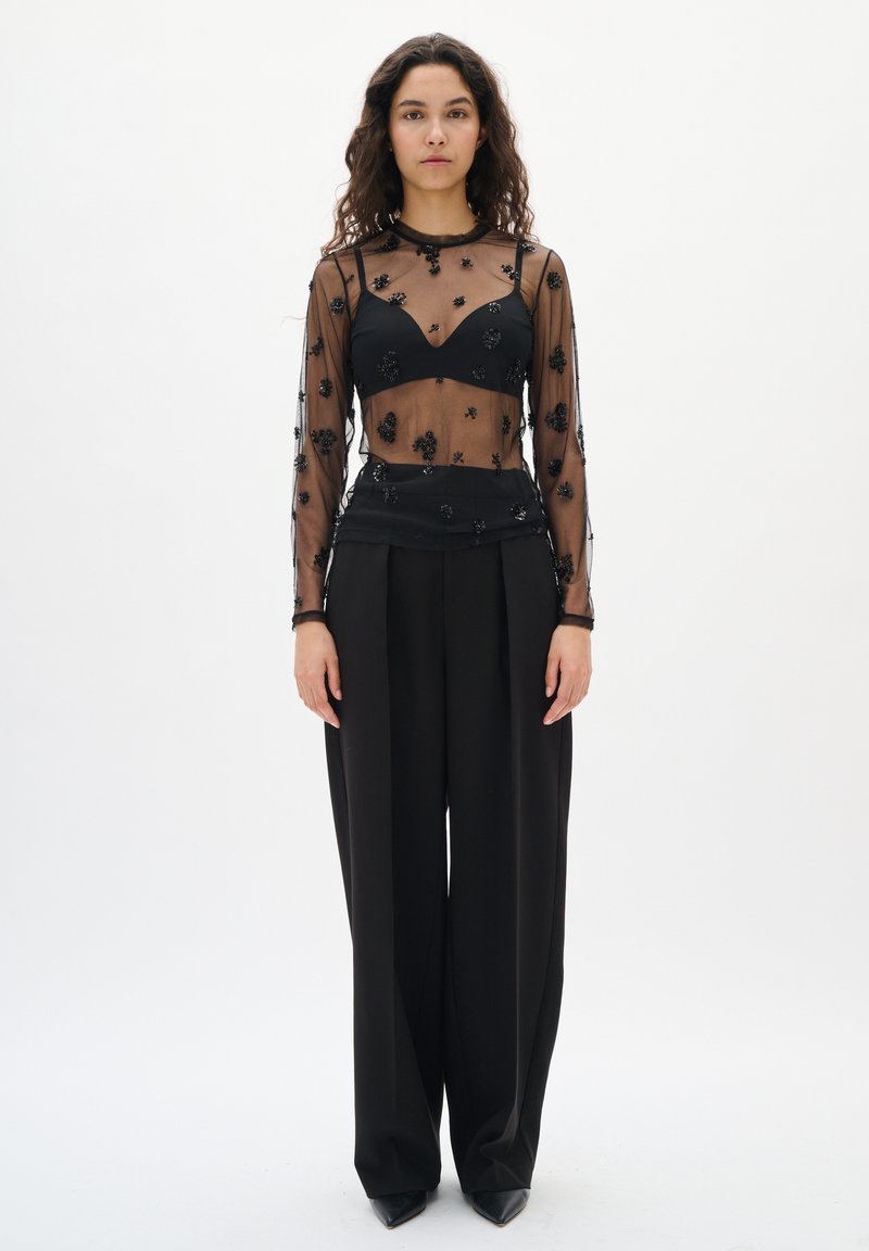 Frau mit lockigem Haar, die ein schwarzes, transparentes Langarmoberteil mit floralem Applikationen, einen schwarzen BH, weite schwarze Hosen und schwarze spitze Schuhe trägt.