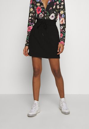 Kvinde iført sort højtaljet nederdel med snøre, blomsterlangærmet top og hvide høje sneakers, stående på ensfarvet baggrund.