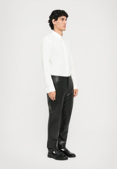 Man wearing white long-sleeve shirt, black textured leather pants, and black loafers, standing against a plain light background.