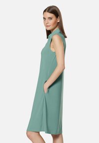 Ärmelloses A-Linien-Kleid aus leicht grünem Stoff, mit Seitentaschen und Kragen, glatter Textur, knielanges Design.
