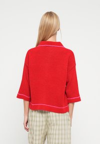 Sarkans adīts džemperis ar īsām piedurknēm, apkakli un rozā detaļām. Valkāts kopā ar zaļiem rūtainiem biksēm. Mīksta struktūra un brīvs piegriezums.