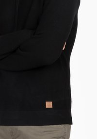 Maglione nero a maglia con texture a coste e maniche lunghe. Presenta un piccolo etichetta in pelle marrone in basso a sinistra, cucita con il nome del marchio.