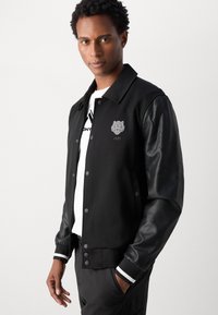 Sort varsity jakke med læderærmer, ribbede manchetter og kant, med et broderet løveemblem og knaplukning foran.
