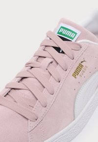Κοντινό πλάνο ενός ροζ sneaker Puma Suede με ροζ κορδόνια, λευκή σόλα και πράσινο λογότυπο Puma στη γλώσσα.