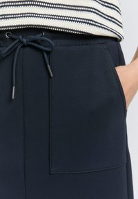 Joggers blu navy con vita regolabile tramite coulisse, dotati di una grande tasca laterale e con una texture di stoffa liscia e opaca. Dettagli includono punte metalliche sulla coulisse.