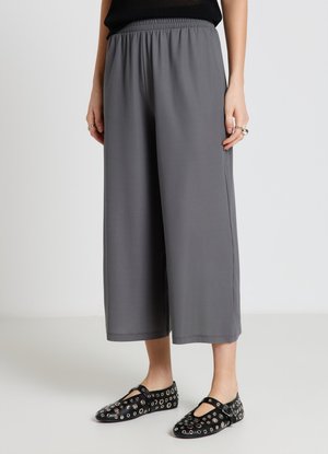 Persona che indossa pantaloni grigi cropped larghi e ballerine Mary Jane nere con dettagli occhiello argento, in piedi su uno sfondo chiaro uniforme.