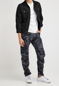 Person klädd i svart jacka, vit t-shirt, mörkgrå kamoflage cargo-byxor, och vita sneakers, stående med ena handen i fickan.