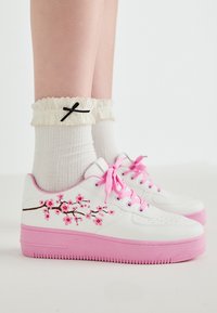 Baskets blanches avec des accents roses, design de cerisier brodé, semelle plate, caoutchouc rose texturé, associées à des chaussettes blanches froncées avec un nœud noir.