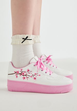 White sneakers with pink accents, embroidered cherry blossom design, flat sole, textured pink rubber, paired with ruffled white socks with a black bow.