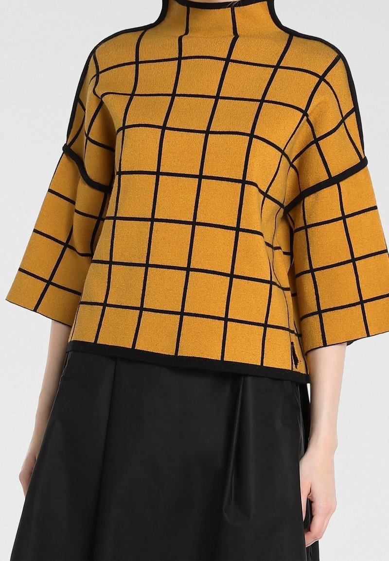Żółty sweter z dzianiny z czarnym wzorem w kratę, z wysokim kołnierzem i krótkimi rękawami. Posiada czarne wykończenie na krawędziach i luźny krój.