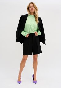 Czarne dopasowane szorty w połączeniu z bluzką w miętowym kolorze, z detalami guzików i marszczonymi mankietami, nałożoną na czarną marynarkę i fioletowe szpilki.