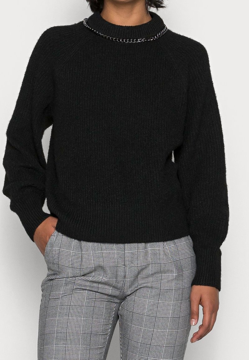 Svart räfflad tröja med rund halsringning, prydd med en metallkedja längs kragen och avslappnade ärmar. Bärs tillsammans med rutiga byxor.