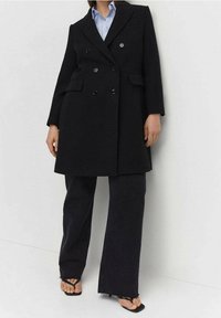 Femme portant un long manteau noir à double boutonnage, un pantalon large noir, une chemise bleue et des sandales à talons noires à bout ouvert.