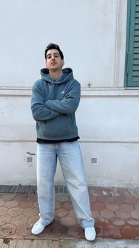 Νέος άντρας με σκούρα μαλλιά που φοράει μια γκρι φούτερ κουκούλα Nike, ανοιχτόχρωμα μπλε τζιν και λευκά αθλητικά παπούτσια, στέκεται με σταυρωμένα τα χέρια μπροστά σε έναν λευκό τοίχο.