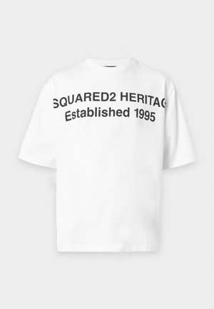 Jeune homme portant un t-shirt blanc oversize avec un texte noir, un jean bleu ample avec des patchs et des chaussures noires, debout sur fond blanc.