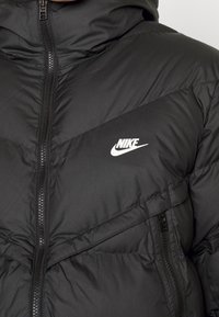 Svart Nike-puffjacka med blixtlås framtill, chevronbroderi och en vit logotyp på vänster bröst. Mjuk, kviltad textur.