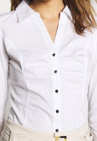 Vit långärmad blus med en figurnära design, med subtila plisseringar och svarta knappar fram. Slät bomullstyg, spetsig krage.