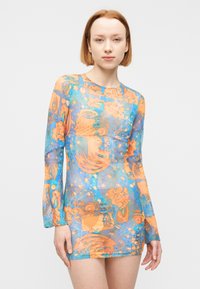 Νέα γυναίκα με κοντά κόκκινα μαλλιά, που φοράει ένα διάφανο, μακρυμάνικο μίνι φόρεμα με αφηρημένο μοτίβο σε πορτοκαλί και μπλε χρώματα.