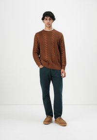 Giovane uomo in piedi contro uno sfondo bianco, indossa un maglione con lavorazione a trecce marrone, pantaloni verde scuro e scarpe allacciate color beige.