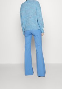 Person klädd i ljusblå utsvängda byxor och en fläckig blå tröja, stående mot en slät vit bakgrund, med ryggen vänd mot kameran.