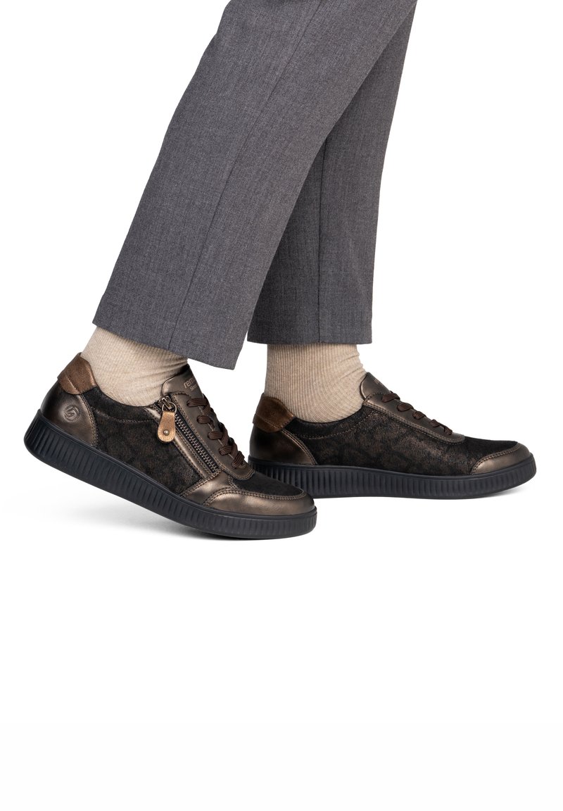 Pieds fermés portant des baskets en cuir et tissu marron foncé avec fermetures éclair latérales, chaussettes beiges et pantalons gris.