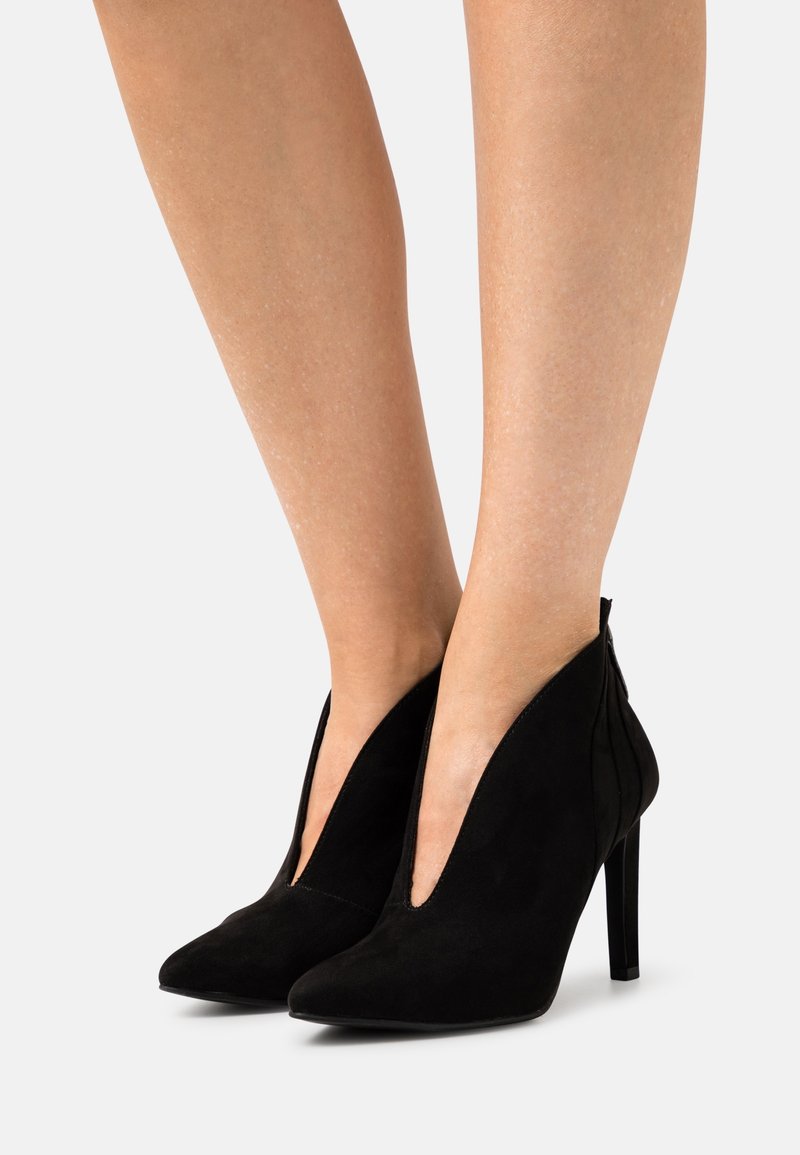 Černé semišové kotníkové boty na vysokém podpatku s špičatou špičkou a bočními výřezem, které mají elegantní, minimalistický design a stiletto podpatek.