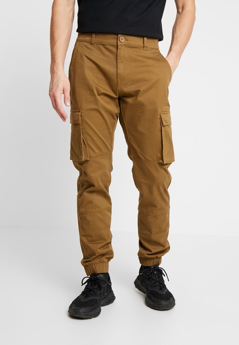 Homme portant un pantalon cargo marron avec des poignets élastiques et des baskets noires, debout avec une main dans la poche devant un fond uni.