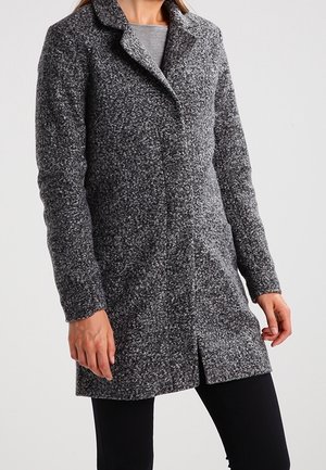 Žena nosící středně dlouhý šedý texturovaný vlněný kabát s dlouhými rukávy, klopovým límcem a skrytým zapínáním v přední části, spárovaný s černými kalhotami.