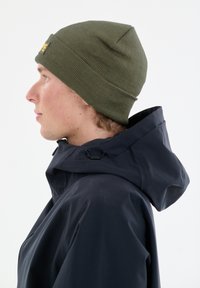 Grüne gestrickte Mütze mit spitzem Oberteil, kombiniert mit einer dunkelblauen Kapuzenjacke, die über einen Kordelzug und sichtbare Nähte verfügt.