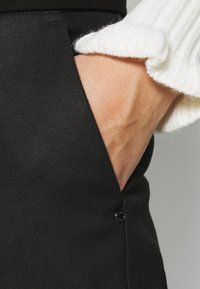 Czarne spodnie materiałowe z boczną kieszenią i ozdobnym guzikiem. Biały prążkowany sweter podkreśla ramię na tle materiału.