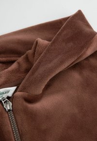 Brązowa miękka kurtka polarowa z widocznym srebrnym zamkiem błyskawicznym i częściowo złożonym kołnierzem, na którym widać fragment białej metki.